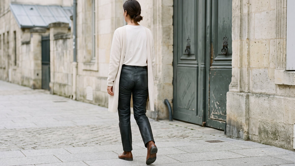 Back view walking along a stone street in straight black leather trousers, a long cream cardigan and brown chelsea boots