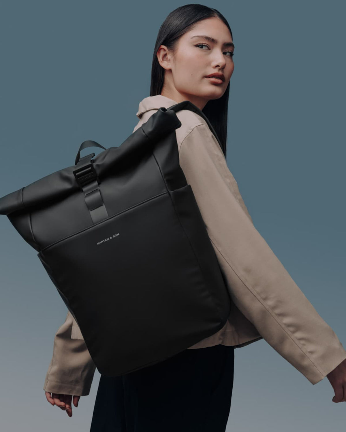 Editorial shot of the all black rolltop backpack carried over one shoulder, beige minimalist styling